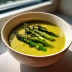 Eine Sch&uuml;ssel mit leuchtend gr&uuml;ner Cremige Spargelsuppe, garniert mit gegrillten gr&uuml;nen Spargelstangen.