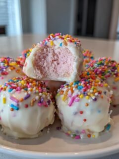 Mehrere knusprige Cotton Candy Cheesecake Bombs, überzogen mit weißer Schokolade und bunten Streuseln, auf einem weißen Teller.