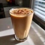 Ein hohes Glas mit einem cremigen, braun-orangen Delicious Apple Pie Smoothie, bestreut mit Zimt.