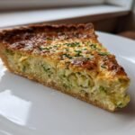 Nahaufnahme eines St&uuml;cks Delicious Artichoke Broad Bean Tart mit goldbrauner Kruste auf einem wei&szlig;en Teller.