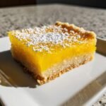 Ein St&uuml;ck saftige Easy Lemon Bars mit dicker Zitronenschicht und Puderzucker auf einem wei&szlig;en Teller.