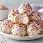 Stapel von Crispy Cotton Candy Cheesecake Bombs, &uuml;berzogen mit wei&szlig;er Glasur und bunten Streuseln, einer angeschnitten.