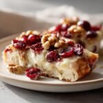 Nahaufnahme eines St&uuml;cks Knuspriges Fladenbrot mit Brie, geschmolzenem K&auml;se, leuchtend roten Cranberrys und Waln&uuml;ssen.