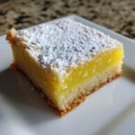Ein einzelnes St&uuml;ck saftige Lemon Bars mit dicker gelber F&uuml;llung und Puderzucker auf einem wei&szlig;en Teller.