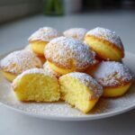 Ein Teller voller gebackener Mini Baby Lemon Impossible Pies, best&auml;ubt mit Puderzucker, einer ist halbiert.