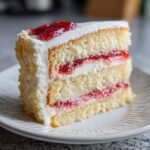 Nahaufnahme eines St&uuml;cks Erdbeer Vanilla Layer Cake mit hellgelbem Biskuit, wei&szlig;er Creme und Erdbeermarmelade.