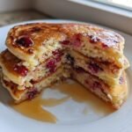 Angeschnittener Stapel fluffiger Cranberry Orange Ricotta Pancakes mit Ahornsirup auf einem wei&szlig;en Teller.
