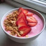 Nahaufnahme eines fertigen Strawberry Smoothie Bowl in einer wei&szlig;en Schale, belegt mit frischen Erdbeerscheiben und knusprigem Granola.