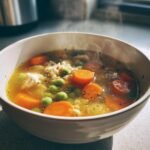 Nahaufnahme einer dampfenden Sch&uuml;ssel mit gesunder H&uuml;hnersuppe mit Gem&uuml;se, Karotten und Erbsen.